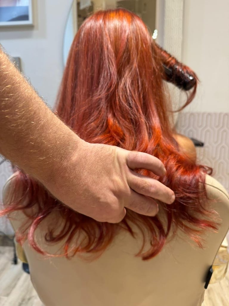 Person with vibrant red-orange hair shown from behind, someones hand touching the hair in an indoor setting