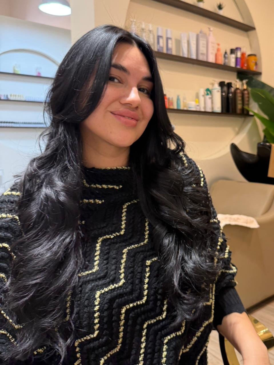 Woman with long black wavy hair wearing a black quilted top with gold accents, smiling indoors with shelves of beauty products in the background