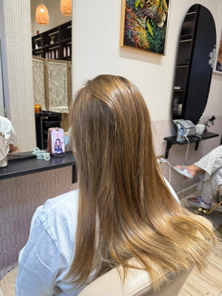 Woman with long, layered blonde hair sitting in a salon chair facing a mirror with black frame and counter display