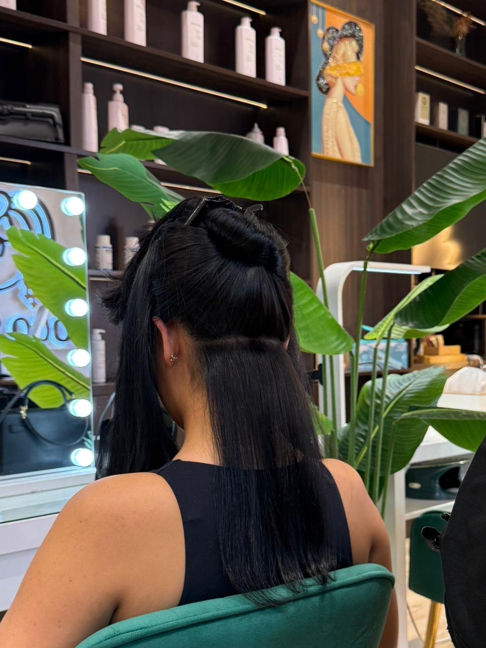 Person with long black hair seated at a salon vanity with lush green plants, illuminated mirror, and shelves with beauty products in background